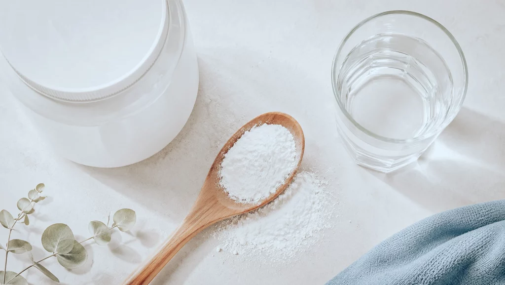 Cuillère en bois remplie de créatine monohydrate en poudre, pot et verre d'eau sur un plan clair