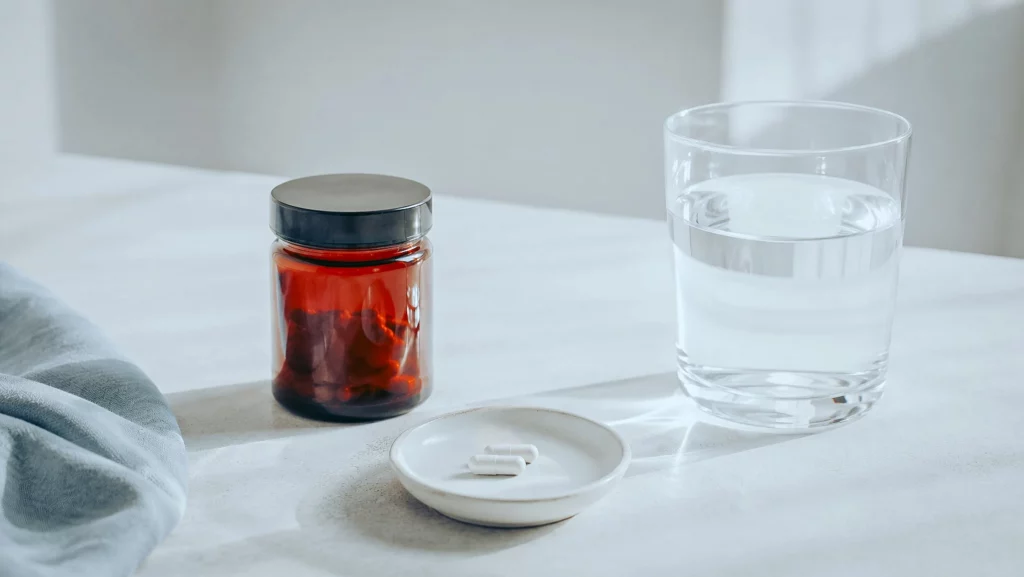 Uma composição sóbria com um frasco, duas cápsulas e um copo de água, evocando uma toma comedida