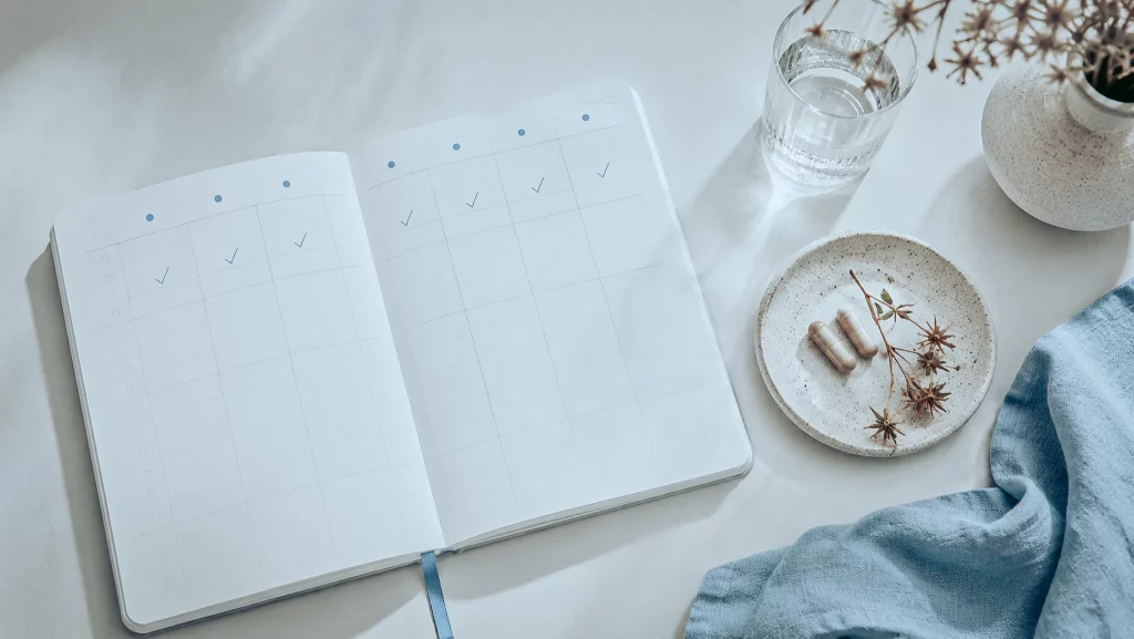 Agenda semanal aberta, um pequeno prato com duas cápsulas e um raminho de Tribulus, evocando regularidade ao longo de várias semanas