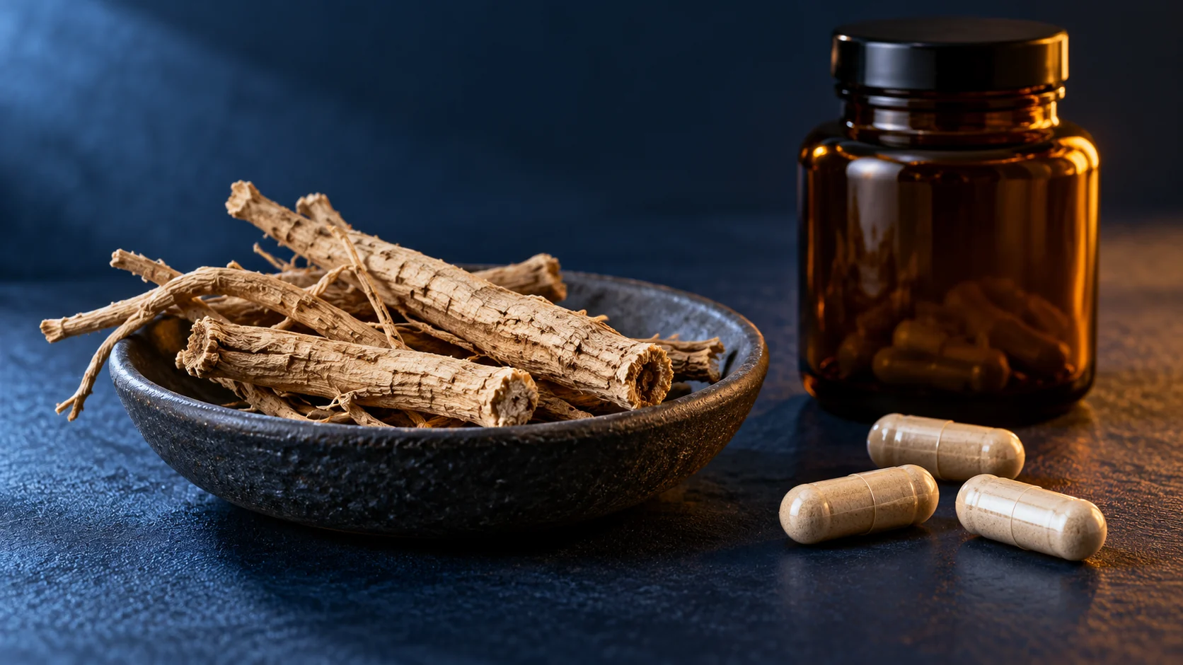 Racines séchées d’ashwagandha dans une coupelle, gélules et flacon ambré sur fond bleu sombre.